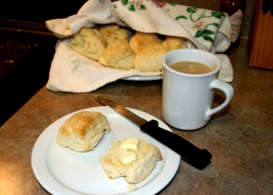 A buttered scone on a plate with a cup of coffee and a large plate of scones in the background